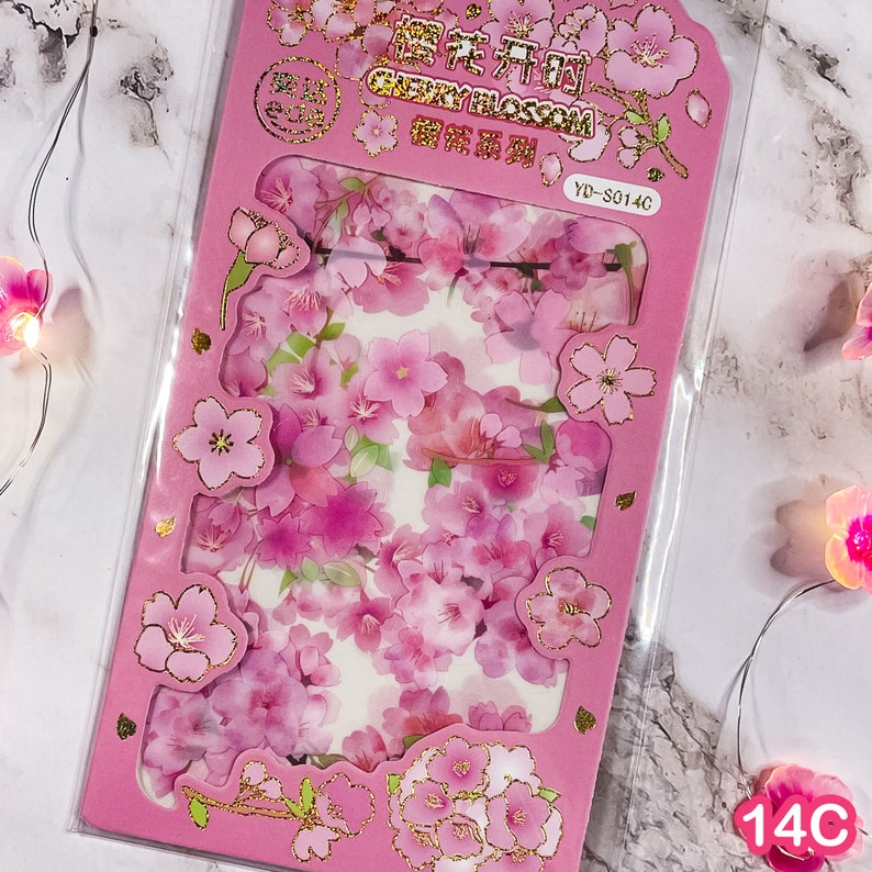 Pu&ograve; includere: Una confezione rosa di adesivi di fiori di ciliegio. Gli adesivi presentano varie tonalit&agrave; di fiori rosa con dettagli dorati. La confezione ha forme ritagliate e testo in oro e nero. Gli adesivi sono in una custodia di plastica trasparente.