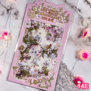 Pu&ograve; includere: Una confezione di adesivi con fiori di ciliegio in una cornice rosa. Gli adesivi presentano fiori bianchi e rosa con foglie verdi. La confezione ha testo dorato e nero. L'angolo in basso a destra ha il numero "14E".