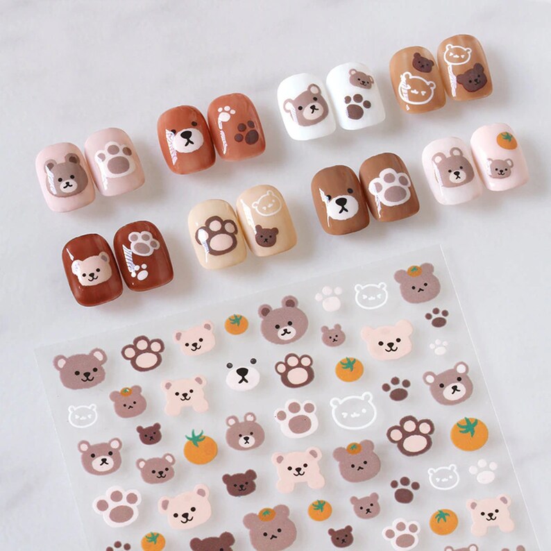 May include: A collection of nail art designs featuring adorable bear and paw print illustrations. The nails are painted in various shades of brown, beige, and white. A sheet of bear and paw print stickers is also visible.