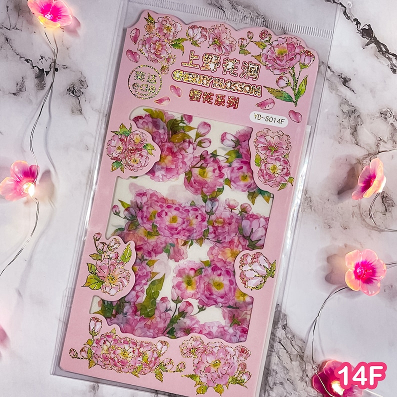 Pu&ograve; includere: Un foglio di adesivi rosa con illustrazioni di fiori di ciliegio in stile acquerello. Il foglio include il testo "CHERRY BLOSSOM" e caratteri cinesi. Gli adesivi sono in una custodia di plastica trasparente. Il foglio ha un bordo rosa con dettagli dorati.