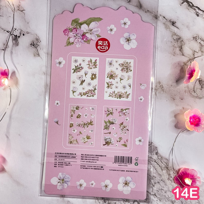 Pu&ograve; includere: Un foglio di adesivi rosa con disegni floreali. Il foglio include vari arrangiamenti di fiori bianchi e viola, con una grande illustrazione floreale in alto. Gli adesivi sono confezionati in una custodia di plastica trasparente.
