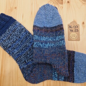 Könnte beinhalten: Ein Paar handgestrickte Socken in Blautönen mit strukturiertem Muster. Die Socken haben einen dunkelblauen Bund und Zehen, mit helleren blauen und braunen Streifen. Ein Etikett mit Größe und Nummer ist angebracht.
