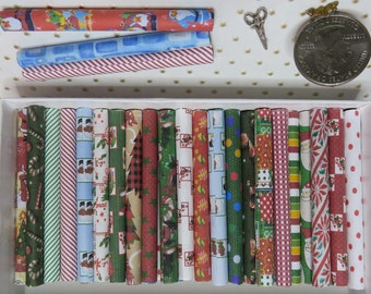 May include: A collection of miniature paper rolls featuring various holiday-themed patterns including Santa, snowmen, candy canes, and Christmas trees, displayed in a white box with a small pair of metal scissors and a coin for scale.