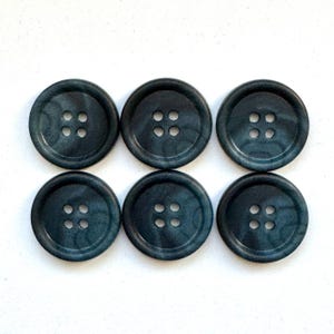 Puede incluir: Seis botones de color verde azulado oscuro con cuatro agujeros cada uno están dispuestos en dos filas de tres sobre un fondo blanco. Los botones tienen un aspecto veteado, con sutiles variaciones de color.