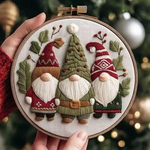 以下が含まれることがあります： 3人の小人が描かれた刺繍のクリスマスフープアート。小人たちは白いひげ、赤と緑の衣装、お祝いの帽子をしています。刺繍は白い背景に施され、緑の葉と赤い実が添えられています。フープは木製です。