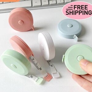 Puede incluir: Una colección de cintas métricas retráctiles en colores pastel, rosa, verde y azul. Cada cinta tiene un botón para retraer la cinta métrica. La imagen también incluye el texto "FREE SHIPPING".