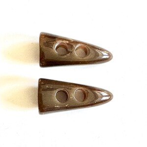 Puede incluir: Dos botones de palanca marrones brillantes. Cada botón tiene forma de triángulo redondeado con dos agujeros para coser. Los botones son de color marrón oscuro liso y parecen estar hechos de un material liso.