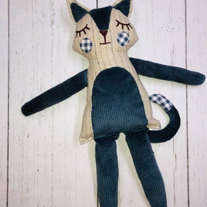 May include: A handmade plush toy cat with a blue corduroy body and a beige head. The cat has black eyes, a brown nose, and a white and blue checkered pattern on its ears and tail.