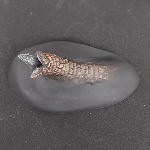 Pode incluir: Uma pequena escultura de cerâmica texturizada, marrom e preta, representando a cabeça de uma criatura emergindo do solo. A criatura tem uma boca aberta com dentes afiados e uma textura escamosa.