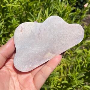 Pode incluir: Uma ferramenta de gua sha em forma de nuvem, feita de pedra natural branca. A ferramenta tem uma superfície lisa e polida e é segurada na mão.