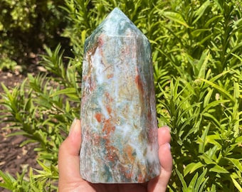 May include: A green, red, and white moss agate crystal point. The crystal is polished and has a smooth, shiny surface. It is held in a hand against a background of green foliage.
