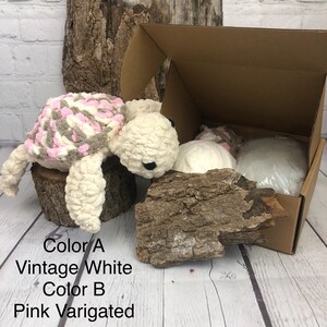 May include: A crocheted turtle plush toy with a cream-colored head and limbs, featuring a shell patterned in pink, white, and grey yarn. The text Color A Vintage White Color B Pink Varigated is printed below the turtle next to a cardboard box.