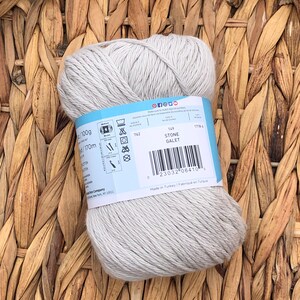 Op de afbeelding: Een bol lichtgrijs katoenen garen met een label met de tekst "Stone Galet" en inclusief wasinstructies en een barcode. Het garen is 100 gram en 170 meter lang.