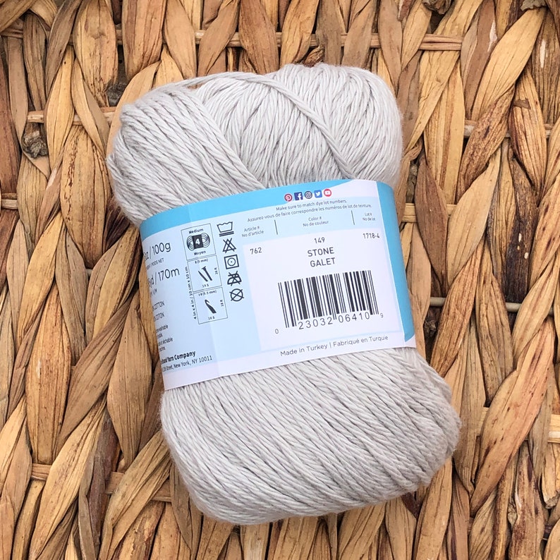 Op de afbeelding: Een bol lichtgrijs katoenen garen met een label met de tekst "Stone Galet" en inclusief wasinstructies en een barcode. Het garen is 100 gram en 170 meter lang.