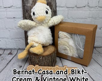 May include: A handmade, cream-colored crochet duckling with a yellow beak and feet, posed on a small wooden slice. The duckling has black eyes and outstretched wings. A brown cardboard box with yarn is visible. Text on the image reads "Bernat Casa and Blkt-Cream & Vintage White Lion Brand FLB-Buttercup."