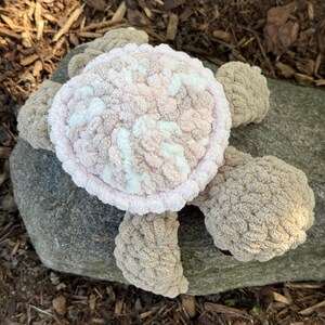 May include: A plush turtle toy crafted from chunky yarn. The shell features a variegated pattern of soft pink, cream, and light brown tones. The head, four flippers, and tail are made from a solid light brown yarn with a textured, looped appearance.