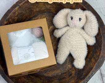 May include: A brown cardboard box with a clear window showing a white and pink yarn kit. The box is sitting on a wooden surface next to a beige plush bunny toy with black eyes and a pink nose. The text "@dezignsbydyan" is written on a wooden tag.