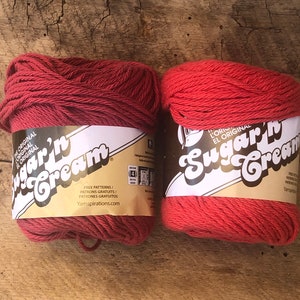 May include: Two spools of red Sugar'n Cream yarn. The label on the spools reads "L'Original El Original Sugar'n Cream" and includes the text "Free Patterns/Patrons Gratuits/Patrones Gratuitos Yarnspirations.com".