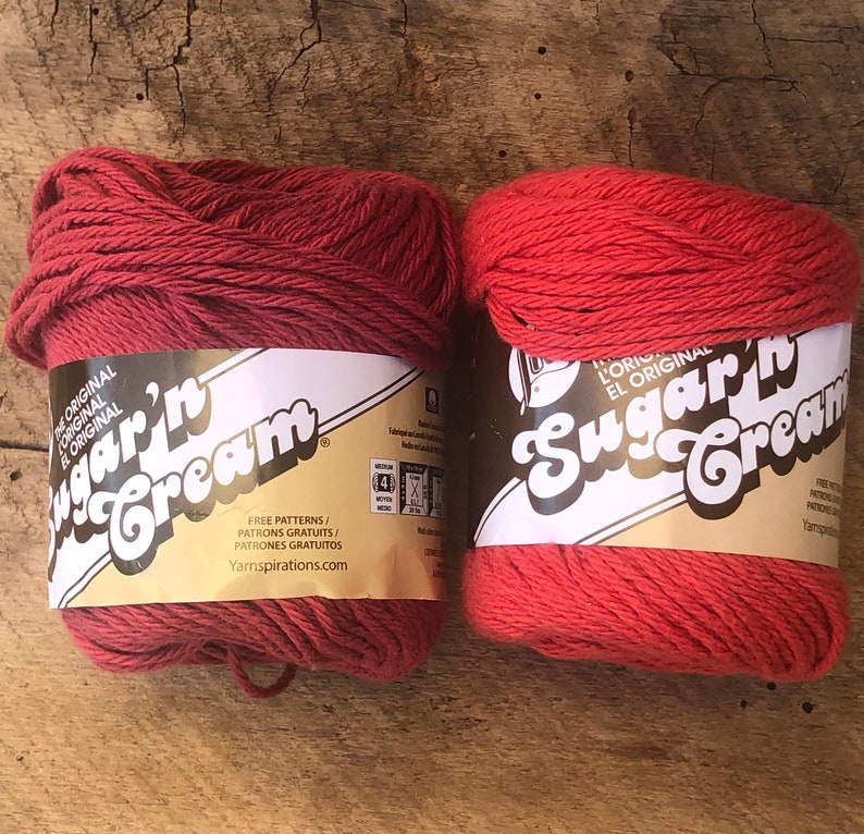 May include: Two spools of red Sugar'n Cream yarn. The label on the spools reads "L'Original El Original Sugar'n Cream" and includes the text "Free Patterns/Patrons Gratuits/Patrones Gratuitos Yarnspirations.com".