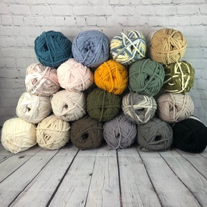 May include: A collection of 18 colourful yarn balls in various shades of blue, green, brown, white, and yellow. The yarn balls are arranged in a pyramid shape on a white brick background.