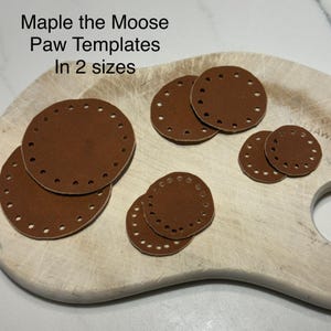May include: Circular brown leather paw templates in two sizes, displayed on a wooden board. Each template has a series of small holes around the perimeter. The image includes the text "Maple the Moose Paw Templates In 2 sizes."