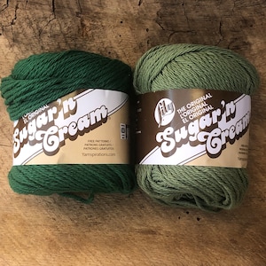 May include: Two skeins of Lily Sugar'n Cream yarn, one in a dark green color and one in a light green color. The yarn is labeled "The Original Lily Sugar'n Cream".