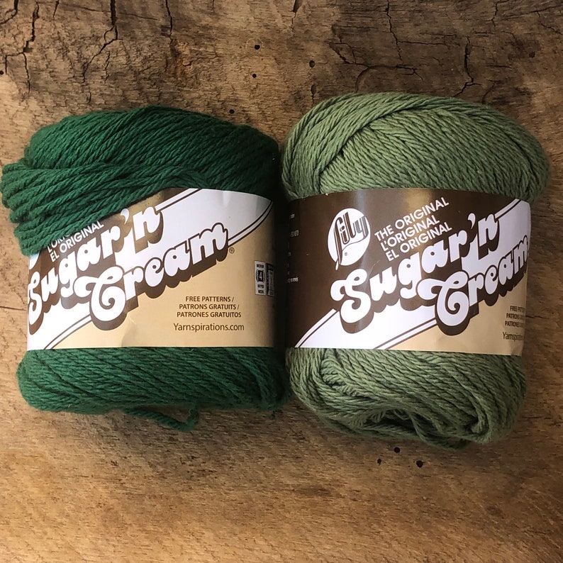 May include: Two skeins of Lily Sugar'n Cream yarn, one in a dark green color and one in a light green color. The yarn is labeled "The Original Lily Sugar'n Cream".