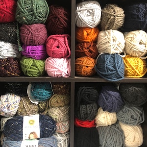 May include: A collection of yarn balls in various colors, including green, purple, pink, and blue, arranged on shelves. The image showcases a variety of textures and shades, perfect for knitting and crochet projects. A label for "Wool Ease" yarn is visible.