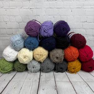 Peut inclure: Une collection de 20 pelotes de laine de différentes couleurs, dont le violet, le bleu, le vert, le gris, le blanc, le jaune, l'orange et le rouge. La laine est disposée en forme de pyramide.