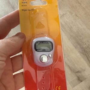 May include: A white finger counter with a digital display and buttons labelled 'Count' and 'Reset'. The counter is attached to a pink wrist strap and packaged on a yellow and red card with the text 'Jixin No 1011 Finger Counter Tally Counter Made in China CE'.
