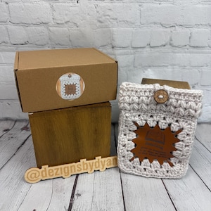 Peut inclure: Un article crocheté à la main de couleur beige, avec un bouton en bois et une étiquette en cuir marron. Il est présenté avec une boîte en bois et une enseigne en bois avec le texte "@designsbydyan".