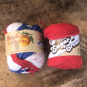 May include: Two skeins of yarn, one red and one white, blue, and red striped. The red skein is labeled "The Original El Original Sugar'n Cream". The striped skein is labeled "Peaches & Creme Stripes, Pistes, Rayas www.peachesandcreme.com"