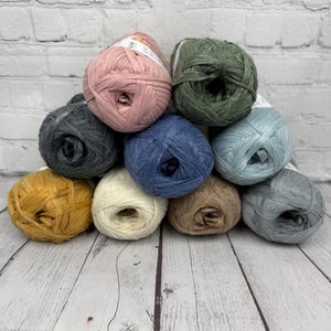 May include: A collection of ten skeins of yarn in various colours, including pink, green, blue, beige, and grey. The yarn is made of a natural fibre, such as cotton or linen, and has a textured, rope-like appearance.