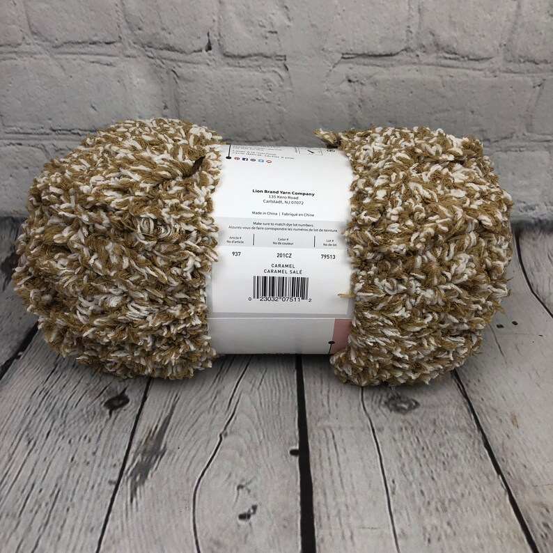 May include: A skein of Lion Brand Yarn Company Caramel Sale yarn in the color 201C2. The yarn is a blend of brown and white and has a fuzzy texture. The label reads "Caramel Sale" and has a barcode.