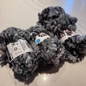 May include: Two skeins of black and white fuzzy yarn with a pink and black stripe. The yarn is labelled "£11.99" and "15 mm".