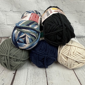 May include: A selection of yarn skeins in shades of blue, green, black, and cream. The yarn is displayed on a light-coloured wooden surface. Some skeins have labels attached.