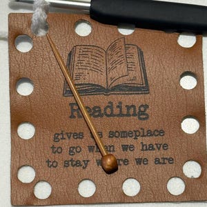 May include: A brown leather bookmark with the word "Reading" and an open book illustration. The bookmark has a series of holes around the edges and the text "gives a someplace to go when we have to stay where we are". A crochet hook and a wooden needle are also present.