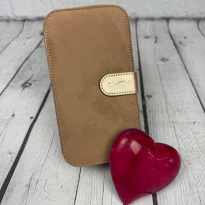 Peut inclure: Un étui de téléphone en daim marron avec un fermoir en métal doré et le mot "Knitter" gravé sur le fermoir. Un objet en forme de cœur rose est au premier plan.
