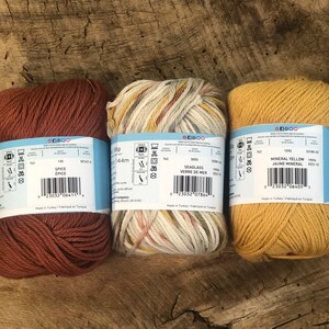 Op de afbeelding: Drie bollen garen in verschillende kleuren: een diep oranje, een beige met bruine en gele strepen en een helder geel. Elke bol heeft een label met de merknaam, de kleurnaam en het gewicht. De labels bevatten ook wasinstructies en barcodes.