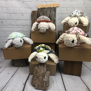May include: Five handmade crochet turtle plushies with soft white yarn heads and limbs. Each turtle features a uniquely coloured shell in patterns of blue, green, brown, pink, and white. The plushies are displayed on brown cardboard boxes and wood.