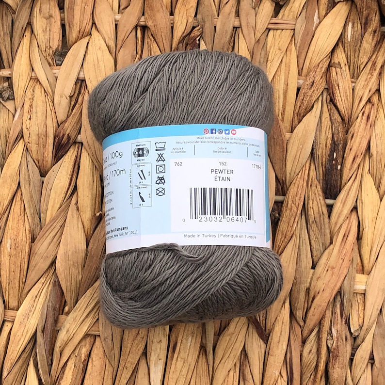 Op de afbeelding: Een bol grijs garen met een label waarop staat "100g / 170m", "Pewter / &Eacute;tain" en "Gemaakt in Turkije / Made in Turkey".