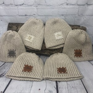 May include: Six beige knitted beanies with brown leather patches. The patches have different text on them, including "Sister", "My Grandkids", "Will", and "Made Grandma Proud".