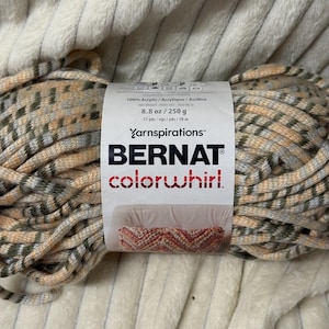 Peut inclure: Pelote de laine Bernat Colorwhirl à motif écossais pêche, gris et vert olive. L'étiquette indique 100% acrylique, 250 g et 70 m. La laine est présentée sur une surface texturée de couleur crème.