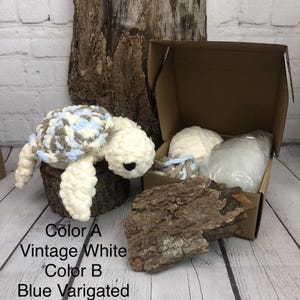 May include: A crochet turtle plush toy with a white body and a variegated shell in shades of blue, white, and brown. Beside it is an open cardboard box containing a ball of white yarn, a small bundle of variegated yarn, and a bag of stuffing.
