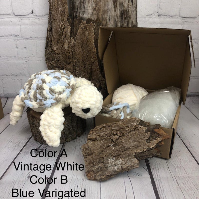 May include: A crochet turtle plush toy with a white body and a variegated shell in shades of blue, white, and brown. Beside it is an open cardboard box containing a ball of white yarn, a small bundle of variegated yarn, and a bag of stuffing.
