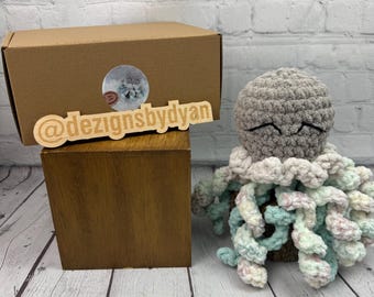 May include: A handmade crochet octopus in shades of gray, white, and pastel blue and pink. The octopus has closed eyes and curly tentacles. The product is next to a brown box and a wooden sign that reads "@dezignsbydyan".