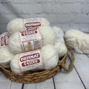 May include: Several white Bernat Haven yarn balls are arranged in a wicker basket. Each ball has a label with the brand name and product information. The yarn is a luxurious alpaca blend, suitable for knitting and crochet.