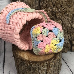 May include: A light pink crochet basket with a blue and green handle, alongside a small, square crochet item. The square item features a pastel colour palette of pink, yellow, blue, and green. Both items are displayed on a wooden surface.