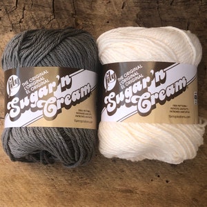 May include: Two skeins of Lily Sugar'n Cream yarn, one in gray and one in white. The yarn is labeled "The Original L'Original El Original Sugar'n Cream" and includes free patterns from Yarnspirations.com.
