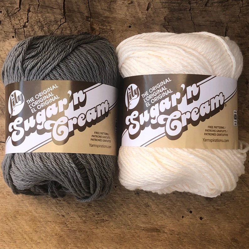 May include: Two skeins of Lily Sugar'n Cream yarn, one in gray and one in white. The yarn is labeled "The Original L'Original El Original Sugar'n Cream" and includes free patterns from Yarnspirations.com.